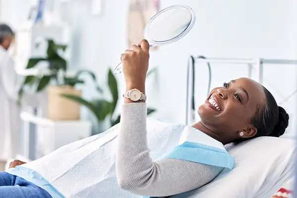 Black woman smiling in hand held mirror after dental cleaning at The Center for Esthetic Dentistry in Grants Pass, OR